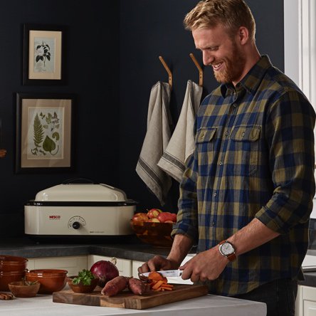 A bearded man who is hosting a holiday dinner party cutting sweet potatoes on a wooden cutting board with a roaster in the background. Starts a blog post about kitchen hacks that make holiday meals easy.